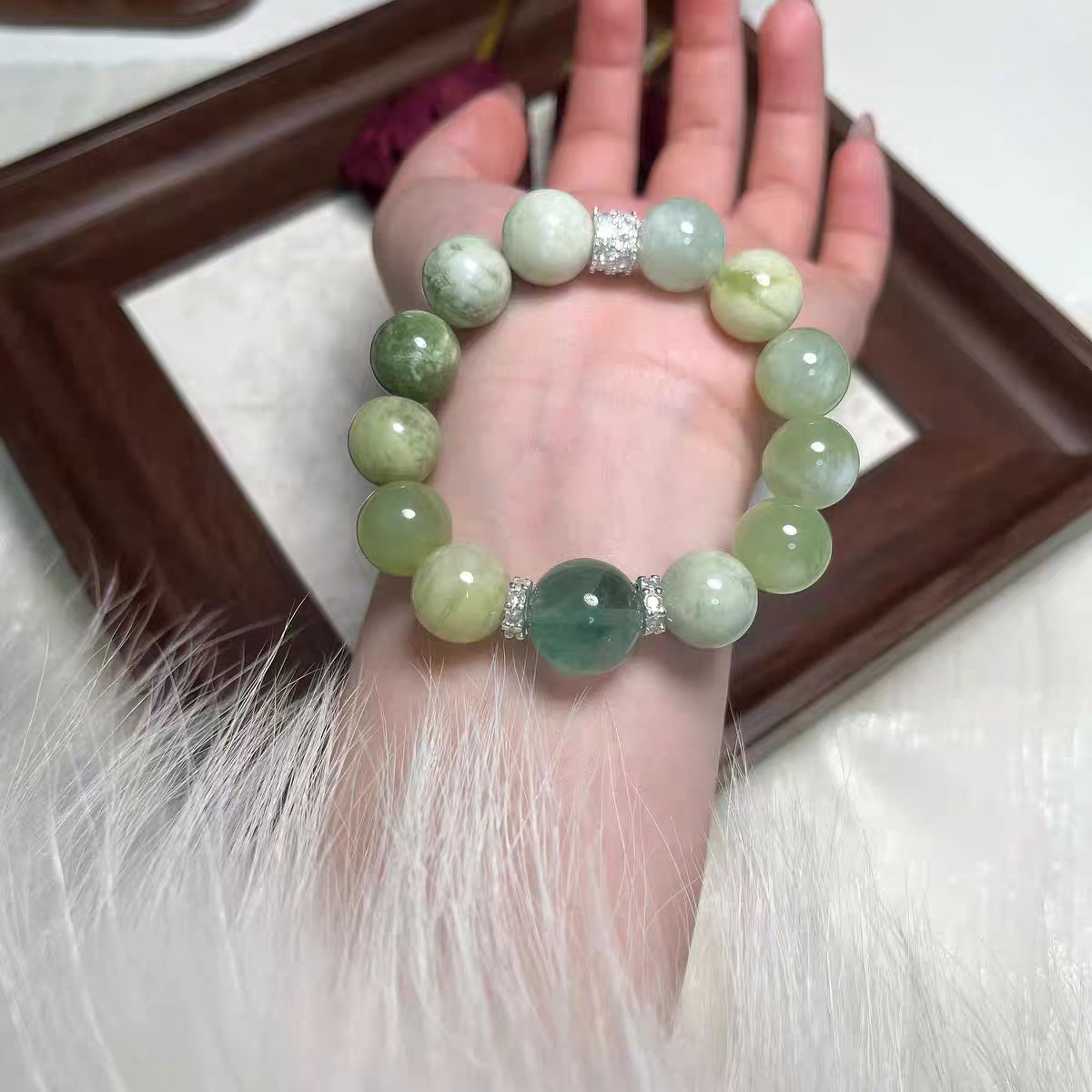 Hand wearing a green beaded bracelet on a white fur surface with a wooden frame in the background